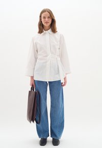 Bela srajca z dolgimi rokavi in pasom na vezanje, v kombinaciji s širokimi modrimi jeans hlačami. Model drži temno rjavo torbo, obut je v črne čevlje.