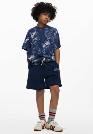 Marineblauw grafisch T-shirt met cartoonfiguren, gecombineerd met marineblauwe terry shorts. Grijze sokken tot aan de kuit met rode strepen en witte sportschoenen.