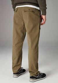 Khaki cargobroek met rechte snit, achterzakken met knoopsluiting en gestikte accenten, gedragen met zwarte sportschoenen.