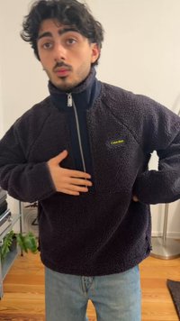 Νέος άνδρας με σκούρα μαλλιά, που φορεί μαύρο φλις μπουφάν Calvin Klein και ανοιχτόχρωμα μπλε τζιν, στέκεται μέσα σε εσωτερικό χώρο με το ένα χέρι στο στήθος.