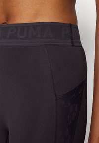 Crne atletske tajice s širokim pojasom na kojem se nalazi utisnuti "PUMA" logo. Glatka tkanina s suptilnom teksturom i detaljima u obliku bočnog džepa.
