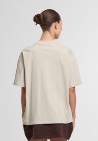 Camiseta oversized de color beige, de manga corta, con cuello redondo, de textura suave, que presenta una etiqueta en la costura lateral y un dobladillo trasero ligeramente alargado.