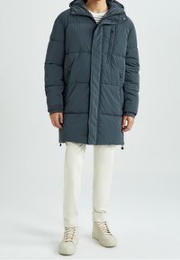 Polstret parka i mørkegrå med hætte, frontlynlås og sidelommer. Har en glat tekstur og elastiske manchetter. Bæres over lyse bukser.