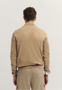 Beżowy sweter z prążkowanym kołnierzem i mankietami, charakteryzujący się gładką fakturą i luźnym krojem. Tło jest neutralne i nierzucające się w oczy.