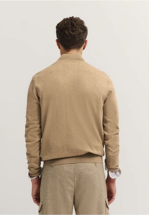 Beige Pullover mit geripptem Kragen und Bündchen, verfügt über eine glatte Textur und einen lässigen Schnitt. Der Hintergrund ist neutral und unauffällig.
