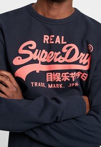 Sudadera azul marino hecha de tela suave con un logo rosa prominente que dice "REAL SuperDry" con texto adicional debajo.