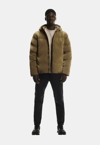 Braune Puffjacke mit Kapuze, featuring einen Reißverschluss vorne, gestepptem Design und gerippten Bündchen, kombiniert mit schwarzen Hosen und Schuhen.