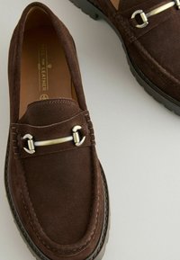 Hnědé suecové loafers s texturovaným povrchem, s kovovým stříbrným akcentem přes nárt a gumovou podrážkou s protiskluzovými gripy.