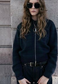 Ciemnozielony i granatowy wełniany sweter z zamkiem, z ryflowanymi ściągaczami, zestawiony z jeansami w ciemnym odcieniu z wysokim stanem i czarnym paskiem. Dodatki obejmują okulary przeciwsłoneczne.