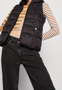 Zwarte puffer vest met een capuchon, voorzien van een rits en drukknoopsluiting, gecombineerd met een gestreept langemouwen shirt en donkergrijze brede broek.