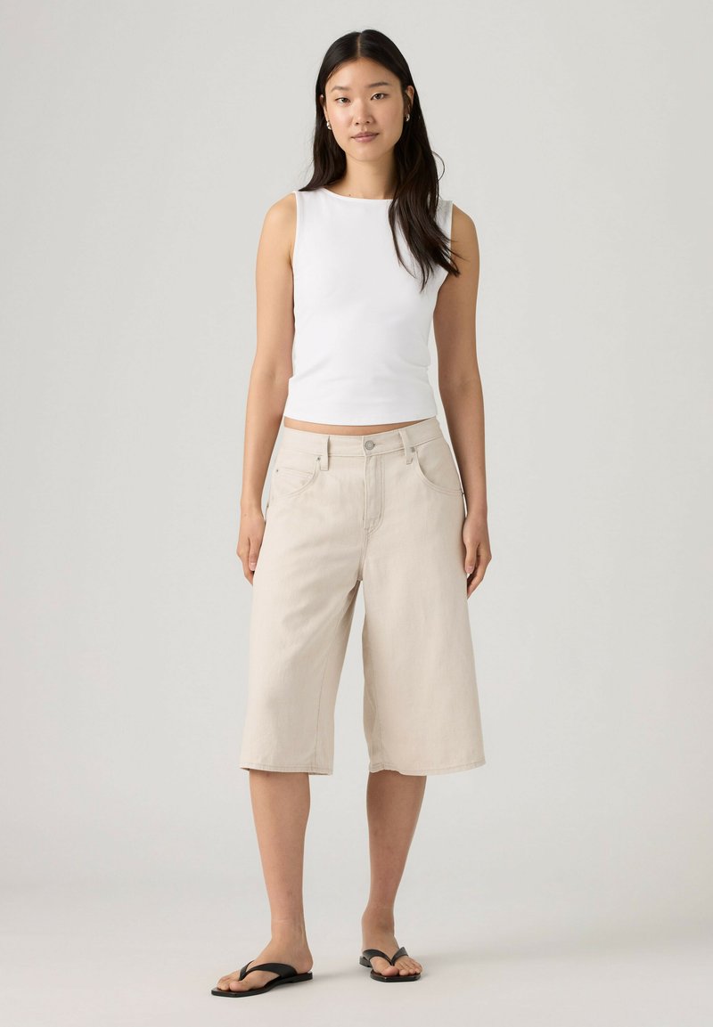 Woman standing wearing a white sleeveless top, beige knee-length wide shorts, and black flip-flops on a neutral background.