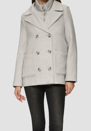 Femme portant un manteau en laine gris clair à double boutonnage avec de gros boutons et des poches avant sur un pantalon sombre.