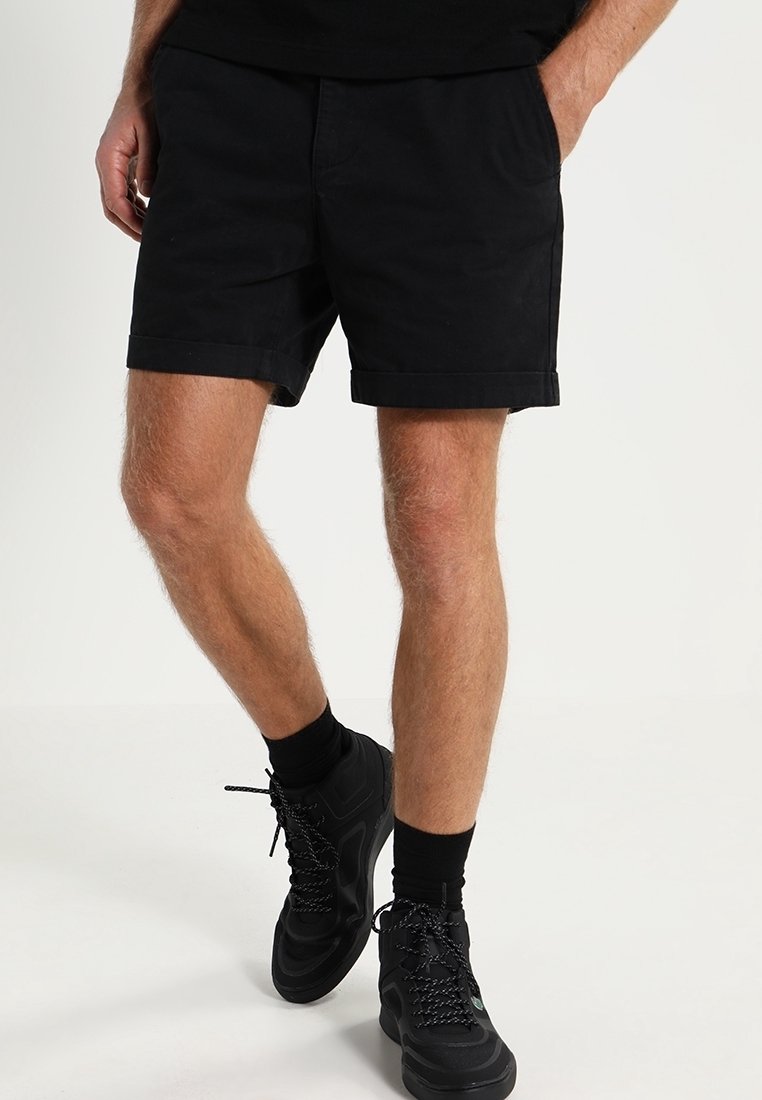 Homme portant un short noir avec un ourlet roulé, des chaussettes noires et des baskets noires, se tenant les mains dans les poches devant un fond uni.