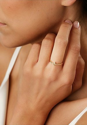Gold ring with a small clear stone, worn on a hand. Smooth finish and slim band, with a light reflection on the gemstone. Skin tone visible.