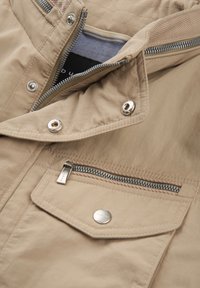 Beige leichte Jacke mit silbernem Zubehör, ausgestattet mit einem Reißverschluss vorne, einer Brusttasche und Klappen-Details. Glatte Textur mit minimaler Naht.
