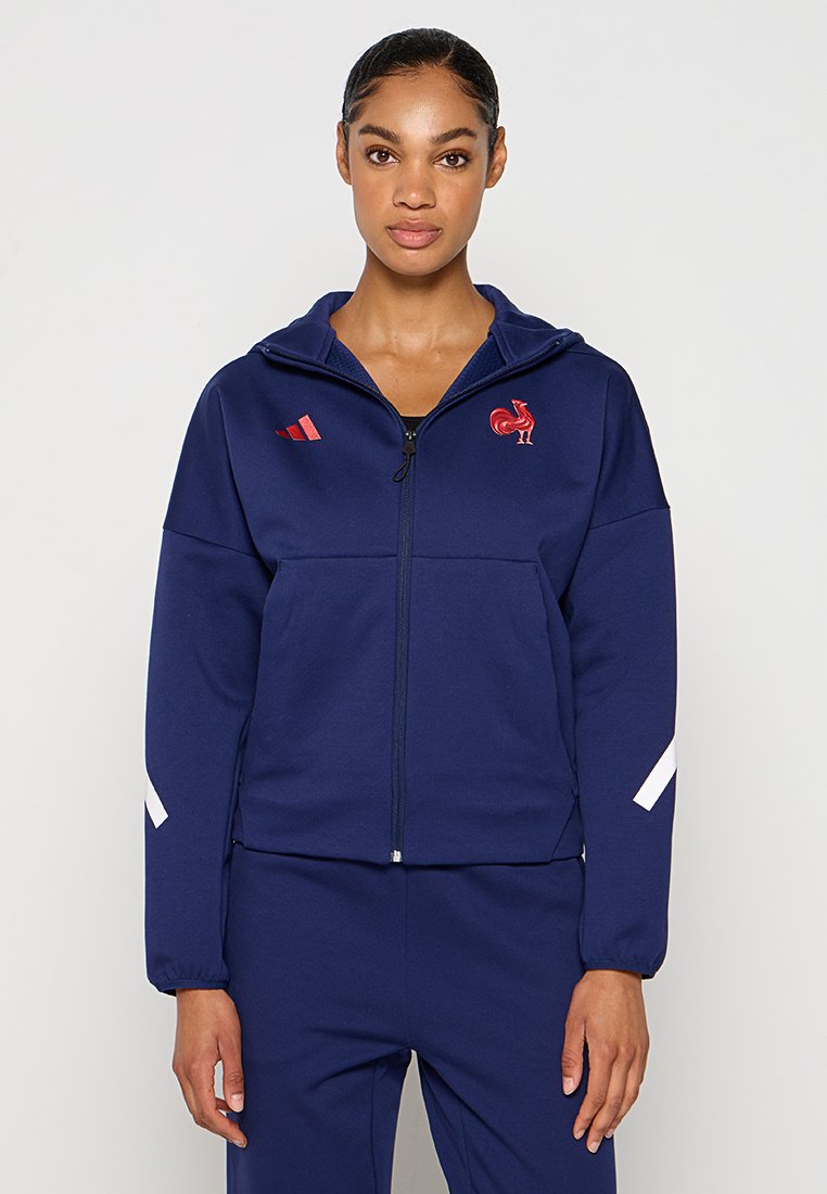 Frau, die einen navyblauen Full-Zip-Hoodie und passende Hose mit dem roten Hahn- und Drei-Streifen-Logo trägt, steht vor einem schlichten hellen Hintergrund.