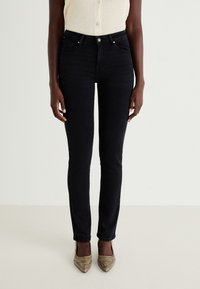 Black skinny jeans made of denim, featuring a high waist, subtle stitching, and front pockets. Worn with a cream top and gold heels.