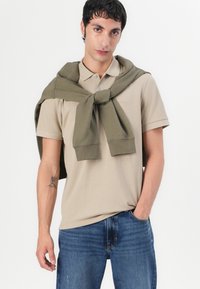 Bež polo majica s kratkim rukavima, koja ima ovratnik i tri gumba, uparena s tamnozelenim puloverom koji je na ramenima. Plave traperice.