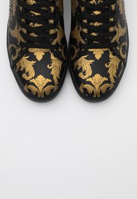 Black sneakers with intricate gold floral patterns, featuring black laces and a rounded toe design. Smooth material and minimal branding visible.
