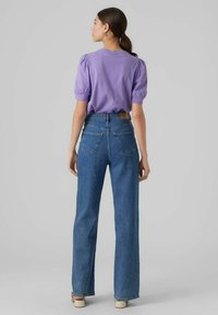 Violet spalvos pūkiniai rankovių marškiniai derinami su aukštu juosmeniu, tiesiomis mėlynomis džinsomis. Džinsai turi užpakalines kišenes ir šviesiai rudos spalvos etiketę ant juosmens.