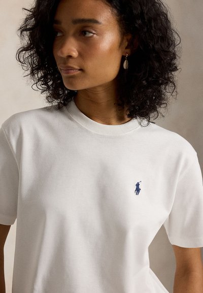 Kobieta o kręconych ciemnych włosach, ubrana w białą koszulkę z krótkim rękawem z małym niebieskim logo gracza polo na piersi, zwrócona lekko w lewo.