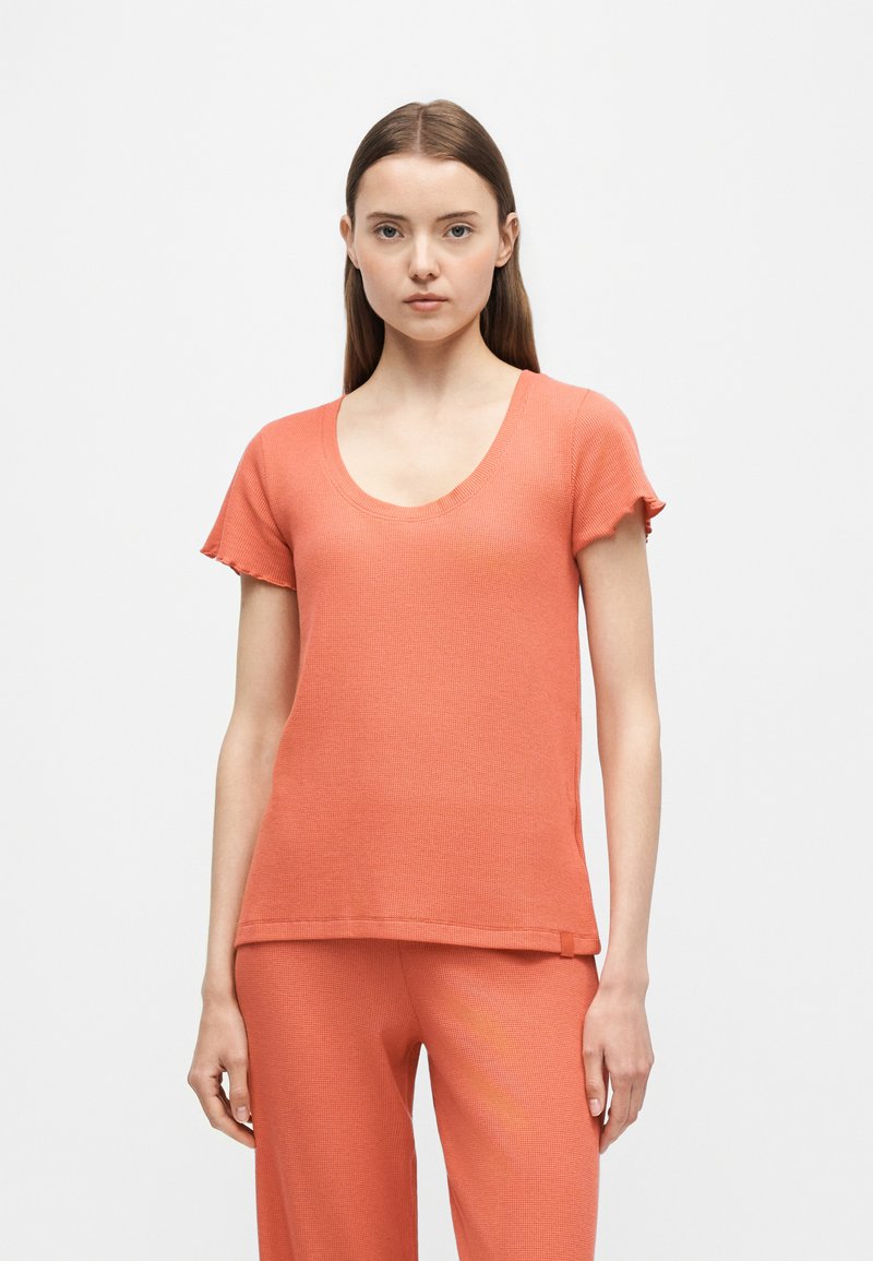 Jeune femme portant un haut court en maille corail avec un col rond et un pantalon en maille corail assorti, debout devant un fond uni.