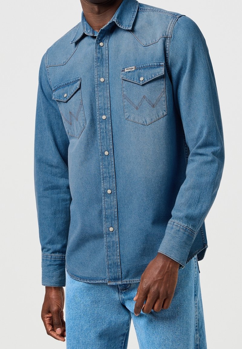 Homme portant une chemise en denim bleu moyen avec des boutons-pression et des poches poitrine à rabat, sur un jean bleu clair.