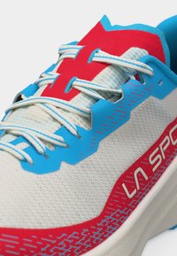 Pantof de alergare cu partea superioară din plasă albă, accente roșii și albastre, șireturi albastre și albe și inscripția "LA SPORT" pe lateral.