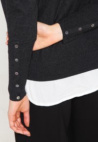 Maglione nero a maglia con texture a coste, con sette bottoni decorativi sulla manica, sovrapposto a un orlo bianco. I pantaloni neri sono parzialmente visibili.