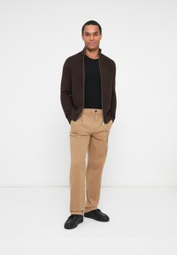 Brun zip-up cardigan over en svart skjorte, sammen med khakifargede bukser og svarte sko. Enkle, rene linjer og nøytral fargepalett.