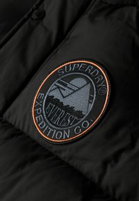 Chaqueta acolchada negra con un parche bordado circular en tonos gris y naranja, que muestra "Superdry Expedition Co." y un diseño de montaña.