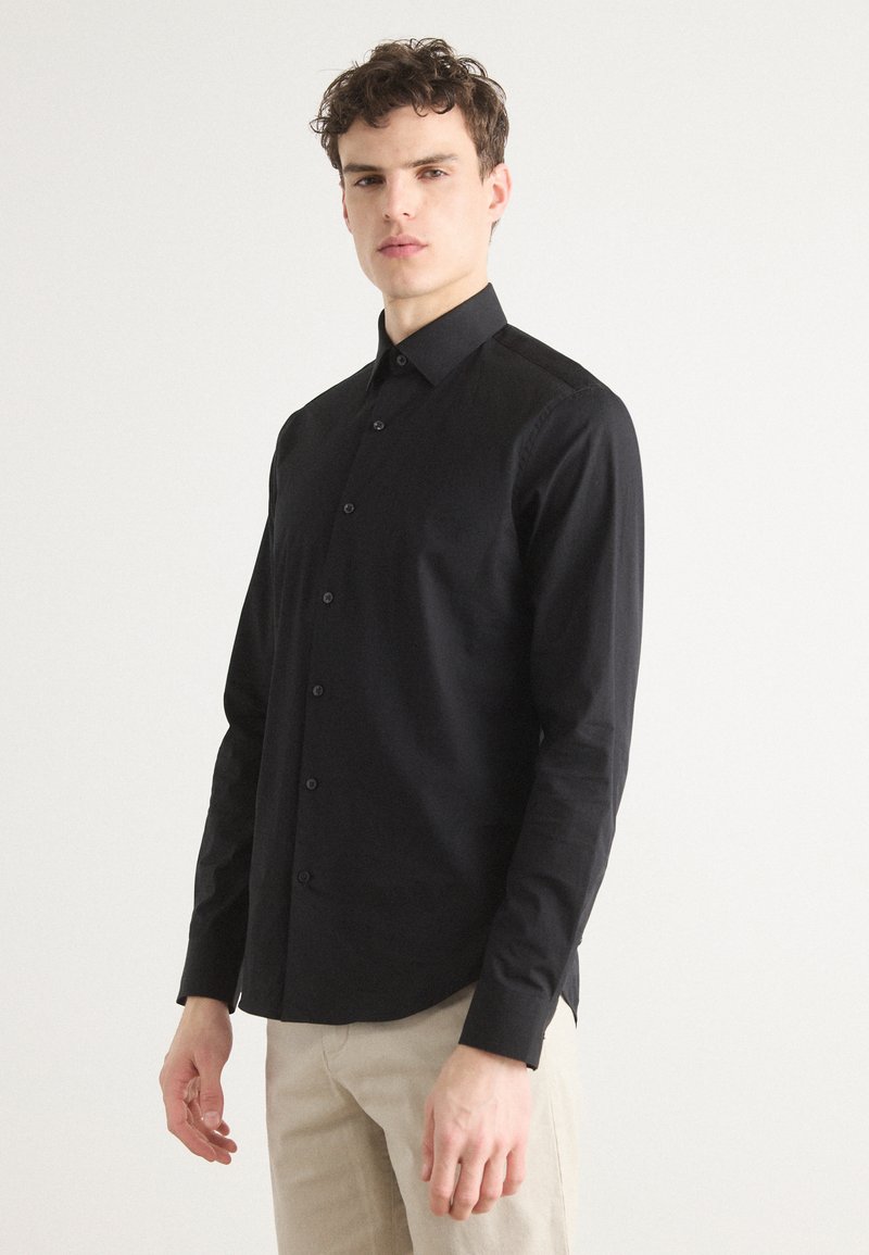 Camisa negra de botones con cuello estructurado y mangas largas. Hecha de una tela suave con una textura sutil. Ajuste entallado con botones visibles.