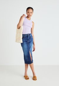 Top in lilla, gonna midi in denim blu scuro con spacco laterale, sandali slide beige e borsa trapuntata chiara. Outfit casual.