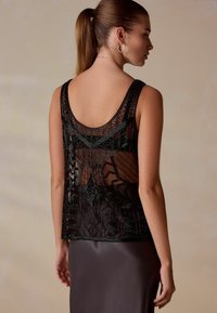 Mustast pärlmutrist topsil keha, millel on geomeetrilise mustriga siidisel tagaküljel. Tal on ümmargune kaelus ja see on kombineeritud satiinhalliga seelikuga.