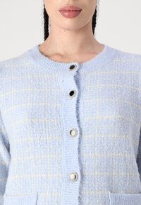 Cardigan bleu clair avec un motif à carreaux texturé, doté de cinq boutons, deux poches avant et des poignets et un col côtelés.