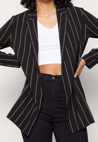 Svart randig kavaj med vita och orange detaljer, med en avslappnad passform, slagkrage och öppen framsidan. Bärs över en vit kort topp.