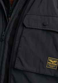 Gros plan d'une poche de veste noire avec un bouton pression et un écusson jaune PME Legend indiquant "American Classic Tested at High Altitude."