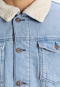 Vaaleansininen farkkutakki, jossa on villakaulus. Ominaisuuksiin kuuluvat metalliset napit, kaksi rintataskua ja ompeluyksityiskohtia.