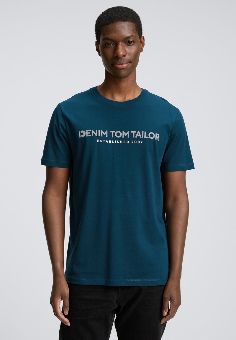 Tirkīza kokvilnas T-krekls ar īsām piedurknēm, apļveida kakla izgriezumu un priekšējo apdruku: "DENIM TOM TAILOR ESTABLISHED 2007" pelēkā krāsā.