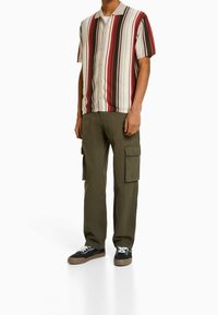 Korte mouwen, knoopshirt met rode, bruine en beige verticale strepen; gecombineerd met olijfgroene cargobroek en zwarte sneakers.