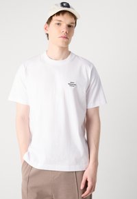 White cotton t-shirt with short sleeves, round neckline, and a small black logo on the chest; paired with beige shorts.
