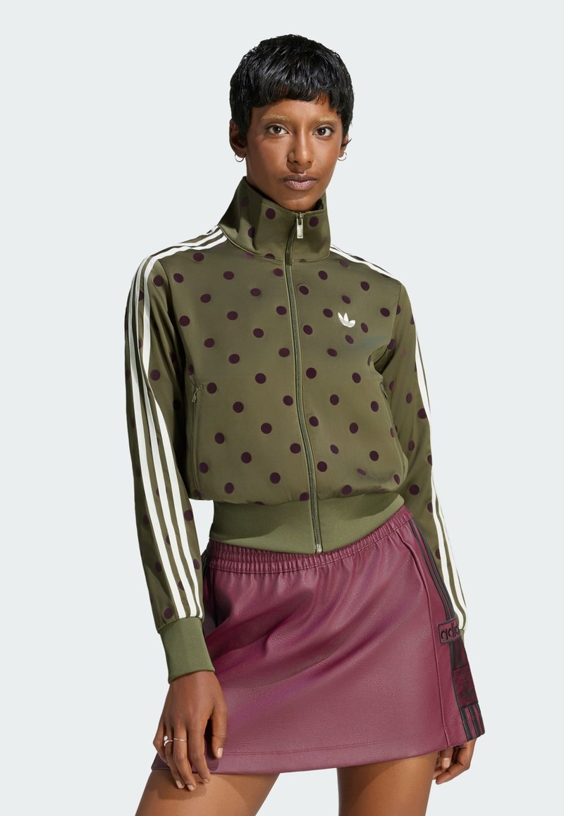 Person wearing an olive green polka dot zip-up jacket with white stripes and a maroon skirt with an Adidas logo patch.