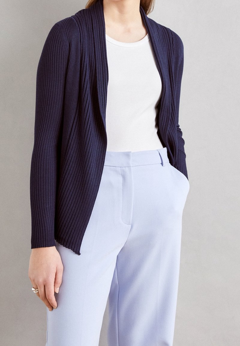 Námořnický žebrovaný cardigan s otevřeným předním dílem, s dlouhými rukávy a uvolněným střihem, nošený přes bílý top a světlé modré kalhoty.