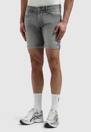 Man met opgerolde grijze denimshorts, witte kousen en zilveren sportieve schoenen, staand tegen een effen achtergrond.