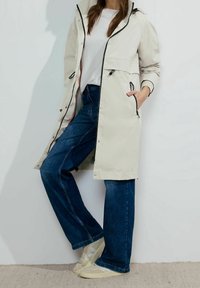 Světlý béžový nepromokavý kabát s kapucí, černými zipy a dvěma bočními kapsami, v kombinaci s tmavě modrými širokými džínami a béžovými teniskami.