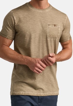Man draagt een beige T-shirt met korte mouwen en een borstzak met knoop, gecombineerd met een donkere broek, de handen voor zich samengevouwen.
