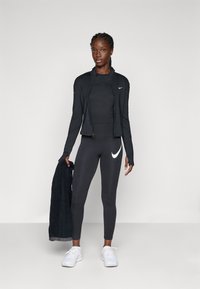 Veste de sport noire avec fermeture éclair, manches longues ; haut noir ajusté ; leggings noirs avec un logo Nike blanc ; baskets blanches ; tenant une serviette foncée.