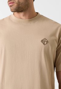 Beżowy bawełniany t-shirt z okrągłym dekoltem i krótkimi rękawami. Na piersi znajduje się subtelne, brązowe haftowane logo. Gładka faktura.