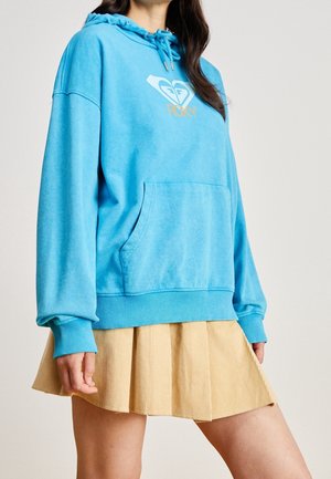 Mujer con una sudadera azul de Roxy con bolsillo delantero y cordones, combinada con una minifalda beige plisada sobre un fondo sencillo.