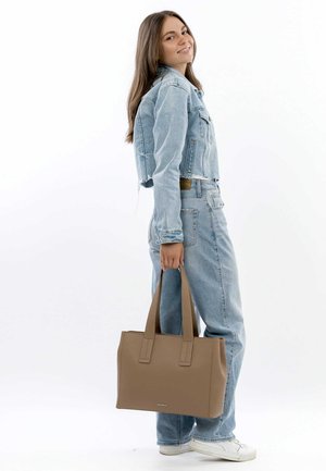 Tan rectangular tote bag with textured surface, dual sturdy handles, and a subtle logo at the front, held by a model in denim outfit.